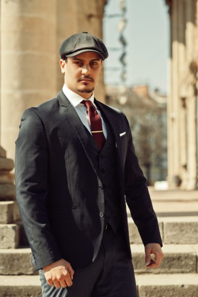 Portrait of retro 1920s english arabian business man wearing dark suit, tie and flat cap near old columns.